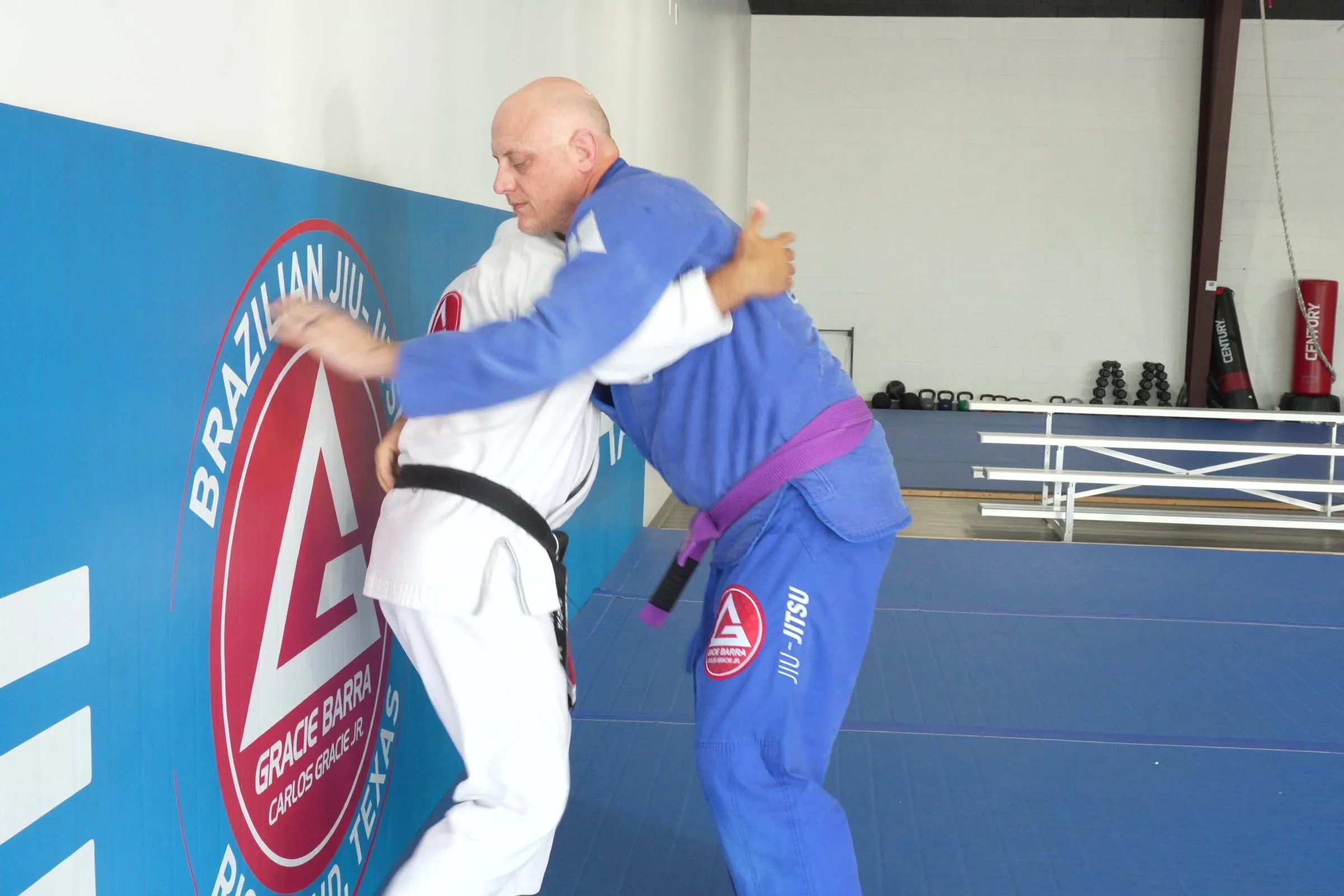 Steve Frassetti in a white gi locked in a standing clinch with a training partner in a blue gi, both men driving into each other with a rack of weights visible behind them.